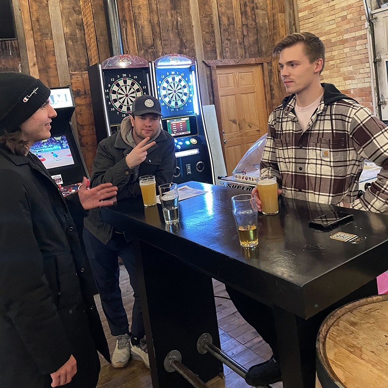 Three members of the Factory talking and standing at a table while drinking beer.